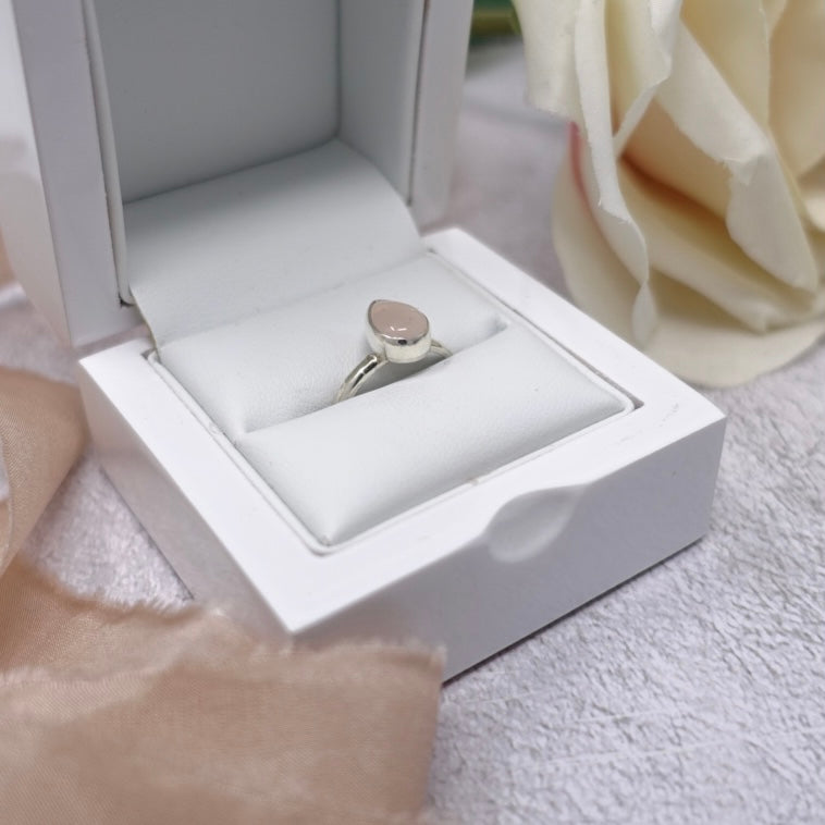 A white ring box sits nestled up against some fraying pink silk ribbon. Inside the box is a beautiful silver and rose quartz teardrop shaped ring