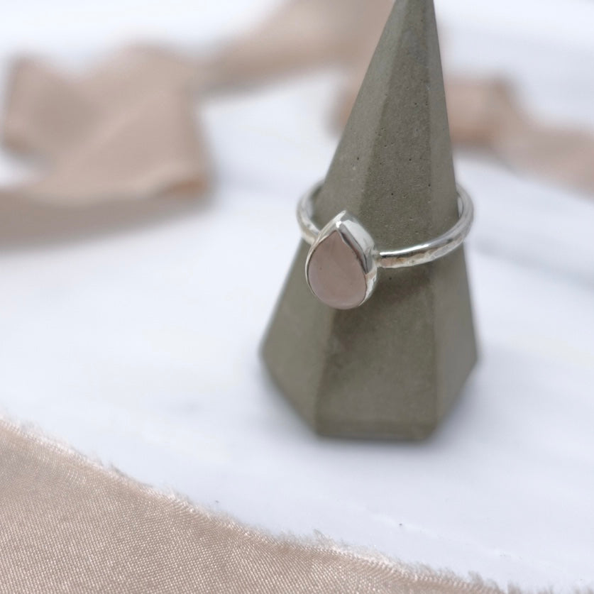 A ring cone sits on a marble background, nestled in amongst some fraying antique pink silk ribbon. On the ring cone sits a soft pink rose quartz teardrop style ring